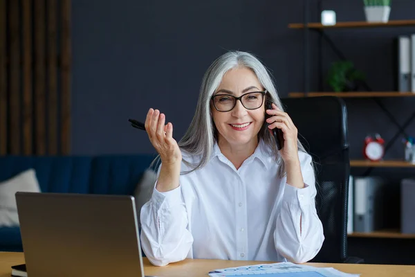 Sonriente mujer de negocios de pelo gris hablando por teléfono en la oficina. Retrato de negocios de una atractiva señora madura con anteojos sentados en el lugar de trabajo. Conversación, negociación —  Fotos de Stock