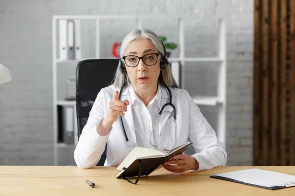 Portrait d'une femme médecin chevelue aux cheveux gris dans son bureau utilisant un ordinateur portable pour le chat vidéo avec une patiente. Consultation en ligne avec le médecin pour les diagnostics et les recommandations de traitement. Concept de télésanté — Photo