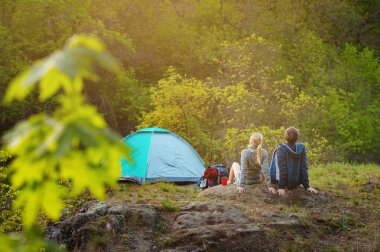 Çiftin seyahat etmek sıcak güneşin çadırda yakınındaki günbatımında dinlenme sırt çantaları ile arkadan görünüm. Seyahat, tatil, tatil ve macera kavramı. Orman dağ manzara arka plan