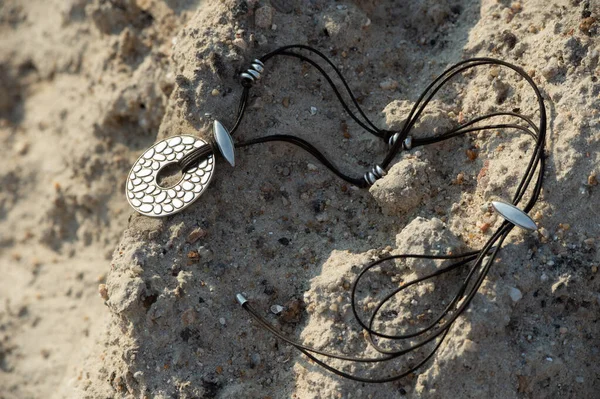 handmade silver pendant on a leather cord lies on the sand by the sea
