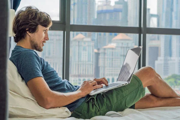 Молодий чоловік працює на ноутбуці в ліжку на фоні панорамного вікна з видом на хмарочоси. Фрілансер, віддалена робота, робота з дому — стокове фото