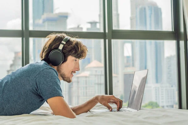 Ung man arbetar på en bärbar dator i sängen på en bakgrund av ett panoramafönster med utsikt över skyskraporna. Använder trådlösa hörlurar. Frilansare, distansarbete, arbeta hemifrån — Stockfoto