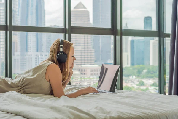 Молода жінка працює на ноутбуці в ліжку на фоні панорамного вікна з видом на хмарочоси. Використовує бездротові навушники. Фрілансер, віддалена робота, робота з дому — стокове фото