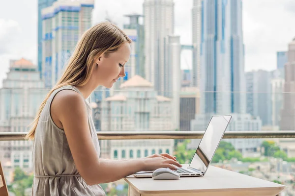 Genç kadın gökdelenler bakan onu balkonda bir dizüstü bilgisayarda çalışıyor. Serbest meslek, uzaktan çalışma, evden çalışma — Stok fotoğraf