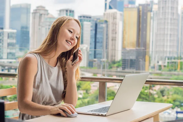 Jonge vrouw is bezig met een laptop op haar balkon met uitzicht op de wolkenkrabbers. Freelancer, externe werkzaamheden, werken vanuit huis — Stockfoto