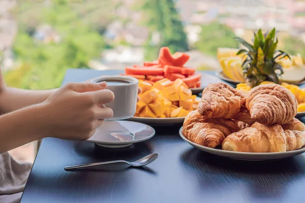 Eine junge Frau frühstückt auf dem Balkon. Frühstückstisch mit Kaffeeobst und Brotcroisant auf einem Balkon vor dem Hintergrund der Großstadt — Stockfoto