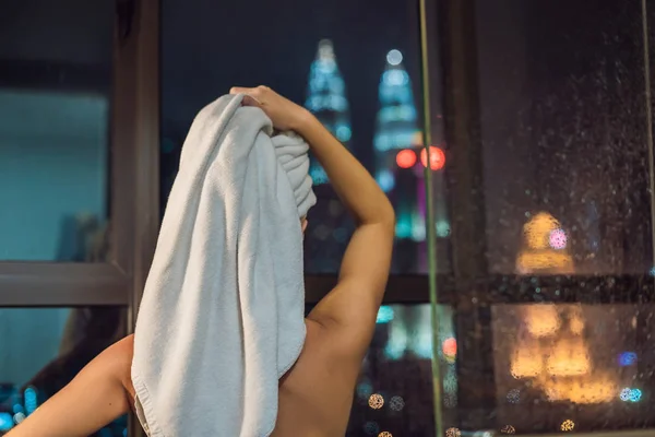 美しい女性は 夜の街のパノラマ ビューとウィンドウの背景色のシャワーの後彼女の髪を拭く — ストック写真
