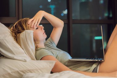 Yatakta dizüstü bilgisayar üzerinde çalışan genç güzel beyaz bir kadın. Gece geç saatlerde büyük şehir manzaralı penceresinin arka plan üzerinde karanlık odada.