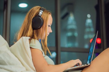 Yatakta dizüstü bilgisayar üzerinde çalışan genç güzel beyaz bir kadın. Gece geç saatlerde büyük şehir manzaralı penceresinin arka plan üzerinde karanlık odada.