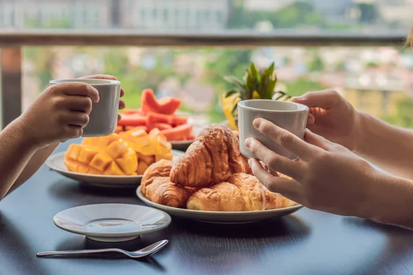 幸せな家族は バルコニーで朝食を有するします コーヒーと朝食用のテーブル 背景には大都市のバルコニーで果物やパンのクロワッサン — ストック写真