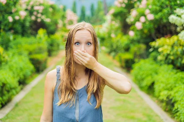 Young woman sneezes in the park against the background of a flowering trees. Allergy to pollen concept.