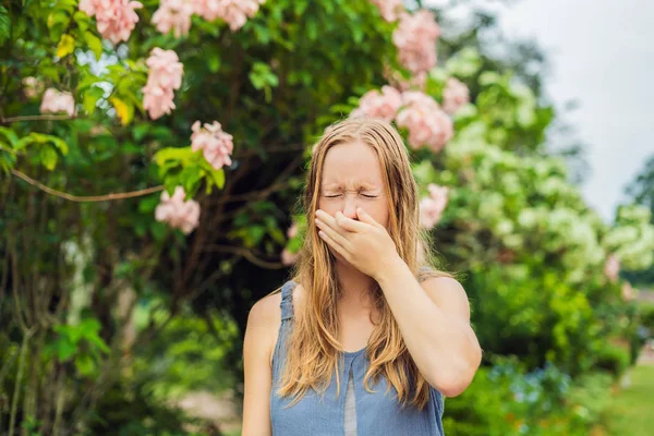 Young woman sneezes in the park against the background of a flowering trees. Allergy to pollen concept.