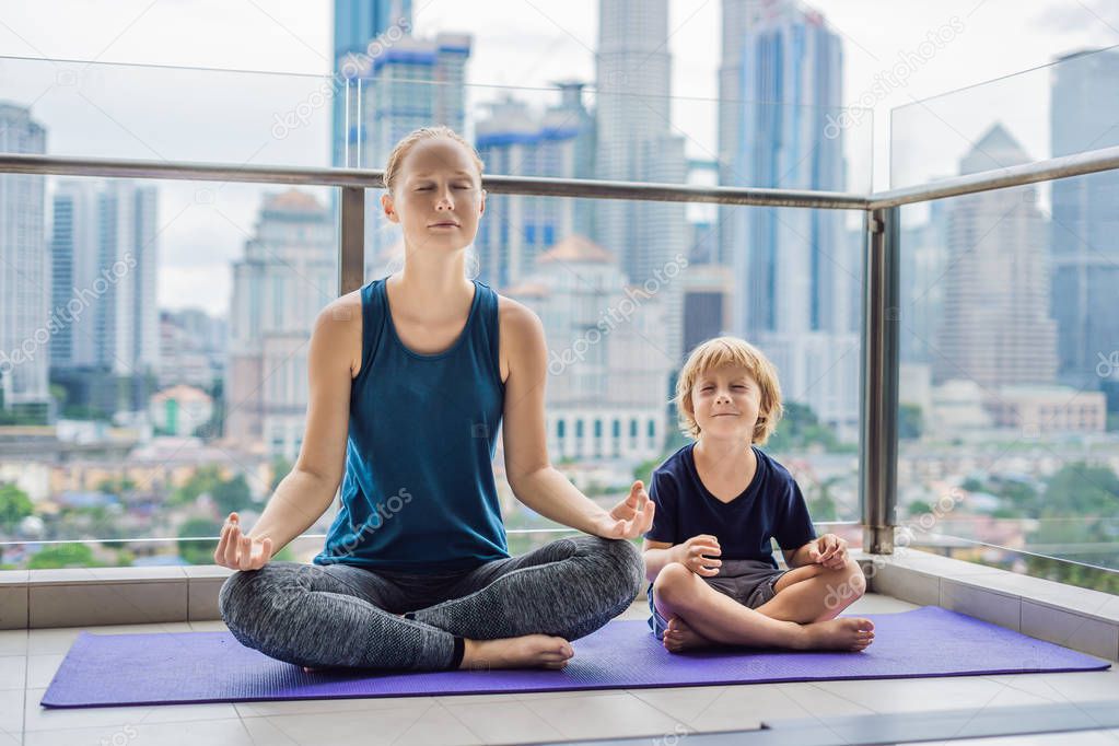 Mom and son are practicing yoga on the balcony in the background of a big city. Sports mom with kid doing morning work-out at home. Mum and child do the exercises together, healthy family lifestyle concept.