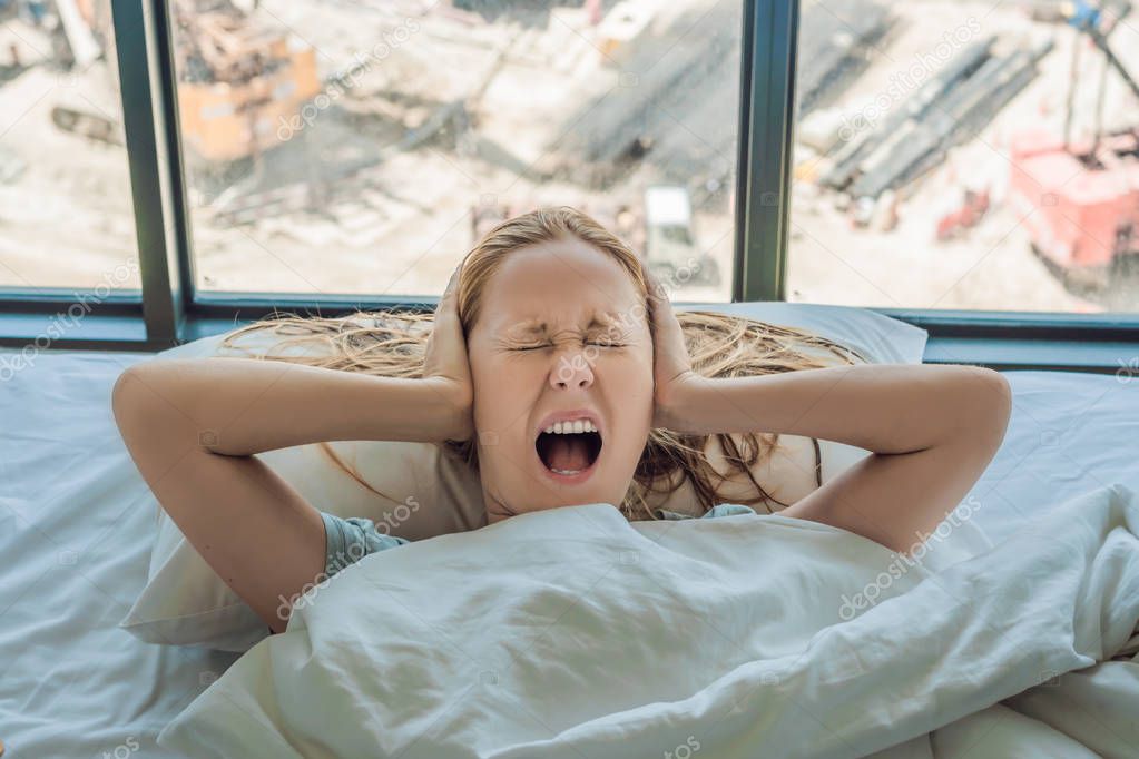 Young woman lying on a bed covered her ears because of the noise. In the window after the bed you can see the construction of a new house.