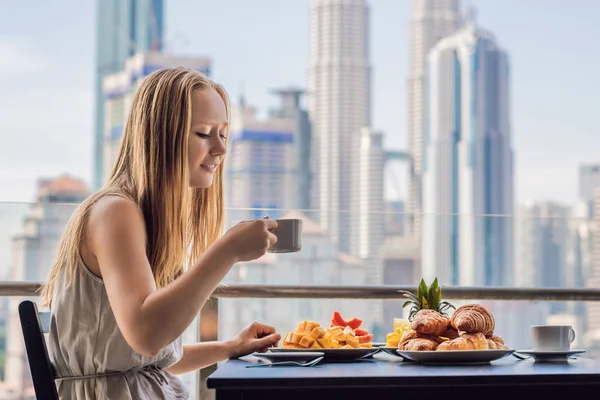 Молода жінка снідає на балконі. Сніданок з кавовими фруктами та хлібом на балконі на тлі великого міста — стокове фото