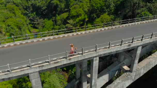 Aerial shot of a woman and her son standing on a tall bridge crossing a canyon with a river on its bottom in tropics. Mother shows something to the boy. — Stock Video