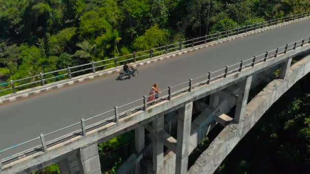 Aerial shot of a woman and her son standing on a tall bridge crossing a canyon with a river on its bottom in tropics. Mother shows something to the boy. — Stock Video