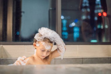 Akşam şehir manzaralı penceresinin arka plan üzerinde uyumaya gitmeden önce akşam banyo küvete oturan mutlu küçük oğlu. Bir banyoda pencere yakınındaki köpük dolu banyo bebek portresi