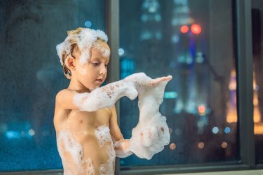 Akşam şehir manzaralı penceresinin arka plan üzerinde uyumaya gitmeden önce akşam banyo küvete oturan mutlu küçük oğlu. Bir banyoda pencere yakınındaki köpük dolu banyo bebek portresi