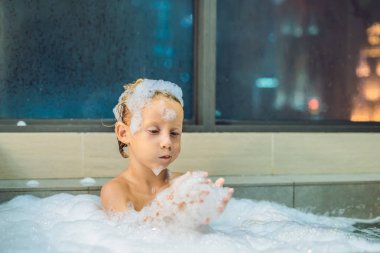 Akşam şehir manzaralı penceresinin arka plan üzerinde uyumaya gitmeden önce akşam banyo küvete oturan mutlu küçük oğlu. Bir banyoda pencere yakınındaki köpük dolu banyo bebek portresi