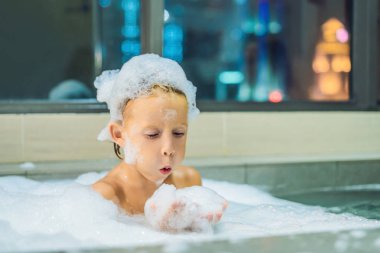 Akşam şehir manzaralı penceresinin arka plan üzerinde uyumaya gitmeden önce akşam banyo küvete oturan mutlu küçük oğlu. Bir banyoda pencere yakınındaki köpük dolu banyo bebek portresi