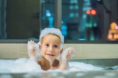 Akşam şehir manzaralı penceresinin arka plan üzerinde uyumaya gitmeden önce akşam banyo küvete oturan mutlu küçük oğlu. Bir banyoda pencere yakınındaki köpük dolu banyo bebek portresi