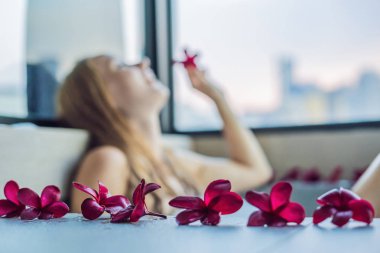 Panoramik pencere bakan gökdelenler ve büyük akşam şehir çerçevede köpük ve frangipani çiçekli banyo genç kadın oturuyor