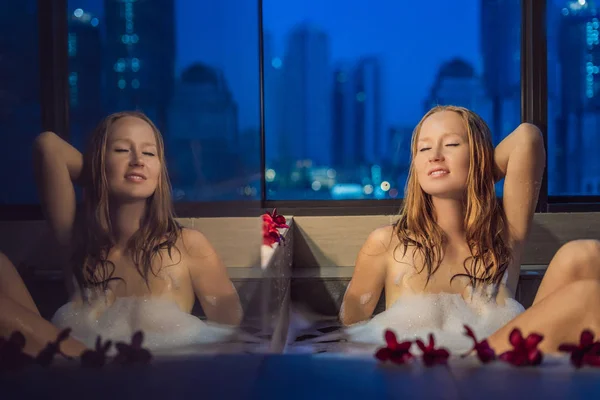 年轻女子坐在浴缸与泡沫和花式花朵反对全景窗口的背景俯瞰摩天大楼和大晚上城市 — 图库照片