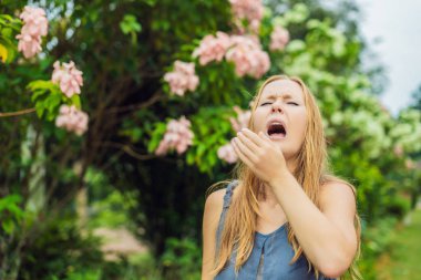 Young woman sneezes in the park against the background of a flowering tree. Allergy to pollen concept. clipart