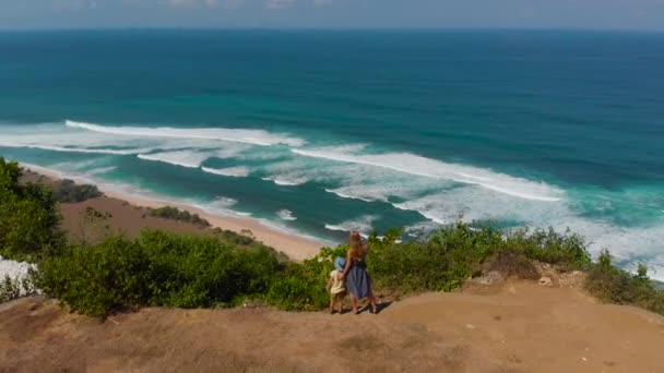 Légi felvétel, egy fiatal nő és a fia, látogatás a távoli tengerparton - Anna Anna - Bali szigetén. Látszó-on egy nyílt óceán előttük egy sziklán álló. — Stock videók