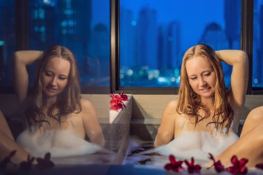 Panoramik pencere bakan gökdelenler ve büyük akşam şehir çerçevede köpük ve frangipani çiçekli banyo genç kadın oturuyor