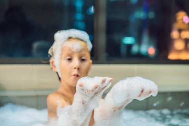 Akşam şehir manzaralı penceresinin arka plan üzerinde uyumaya gitmeden önce akşam banyo küvete oturan mutlu küçük oğlu. Bir banyoda pencere yakınındaki köpük dolu banyo bebek portresi
