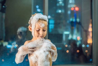 Akşam şehir manzaralı penceresinin arka plan üzerinde uyumaya gitmeden önce akşam banyo küvete oturan mutlu küçük oğlu. Bir banyoda pencere yakınındaki köpük dolu banyo bebek portresi