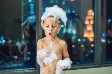Akşam şehir manzaralı penceresinin arka plan üzerinde uyumaya gitmeden önce akşam banyo küvete oturan mutlu küçük oğlu. Bir banyoda pencere yakınındaki köpük dolu banyo bebek portresi