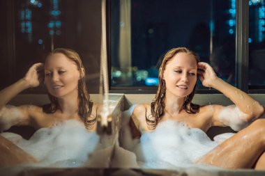 Panoramik pencere bakan gökdelenler ve büyük akşam şehir çerçevede köpük ve frangipani çiçekli banyo genç kadın oturuyor