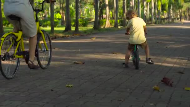 Standbild einer jungen Frau und ihres kleinen Sohnes auf einem Fahrrad und Laufrad in einem tropischen Park — Stockvideo