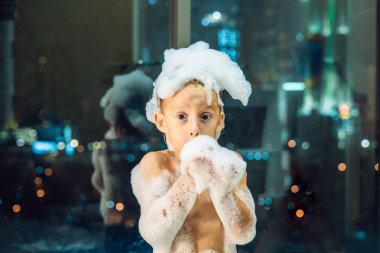 Akşam şehir manzaralı penceresinin arka plan üzerinde uyumaya gitmeden önce akşam banyo küvete oturan mutlu küçük oğlu. Bir banyoda pencere yakınındaki köpük dolu banyo bebek portresi