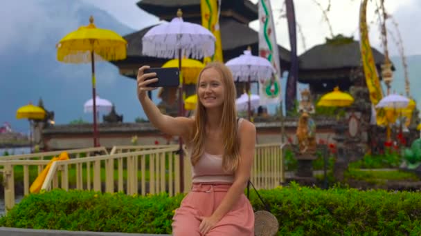 Slowmption tiro de uma jovem mulher fazendo selfie sentado na frente de um templo Pura Ulun Danu no lago Bratan em Bali, Indonésia — Vídeo de Stock