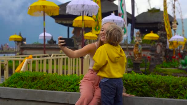 Slowmption tiro de uma jovem mulher e seu filho tomando selfie sentado na frente de um templo Pura Ulun Danu no lago Bratan em Bali, Indonésia — Vídeo de Stock