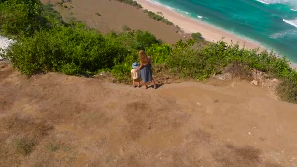 Slowmotion εναέριο πλάνο του μια νεαρή γυναίκα και το γιο που επισκέπτονται μια απομακρυσμένη παραλία - nyang nyang-σχετικά με το νησί του Μπαλί. Στέκεται πάνω σε ένα βράχο που βλέπουν σε μια ανοικτή ωκεανό μπροστά τους. — Αρχείο Βίντεο