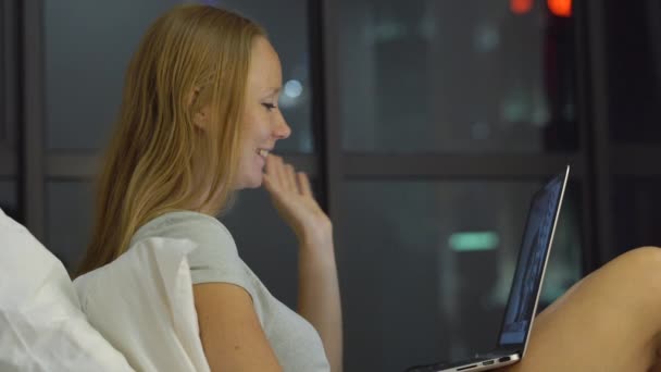 Mladá žena s conveersation pomocí přenosného počítače sedí na posteli s siluety skyscrappers na pozadí — Stock video