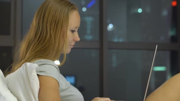 Jonge vrouw met behulp van een laptop zitting op haar bed met silhouetten van skyscrappers op een achtergrondkleur — Stockvideo