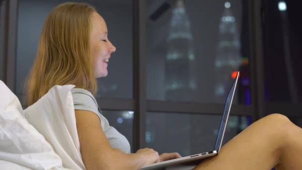 Jeune femme ayant une conversation à l'aide d'un ordinateur portable assis sur son lit avec des silhouettes de gratte-ciel à un backgound — Video