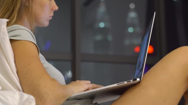 Mladá žena pracuje, pomocí přenosného počítače sedí na posteli s siluety skyscrappers na pozadí — Stock video