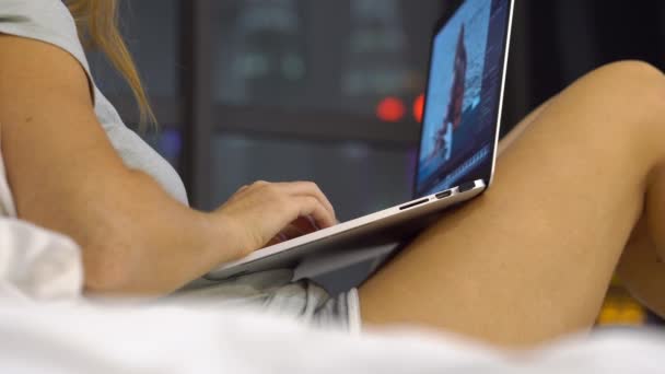 Jovem mulher trabalhando usando um laptop sentado em sua cama com silhuetas de arranha-céus em um backgound — Vídeo de Stock