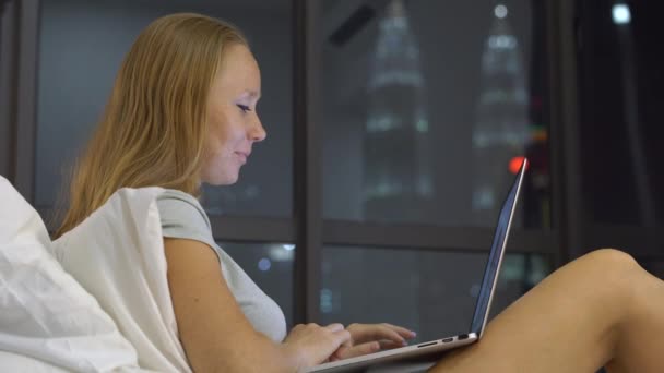 Giovane donna che ha una conversazione utilizzando un computer portatile seduto sul suo letto con sagome di grattacieli a un backgound — Video Stock