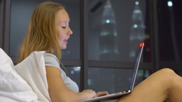 Jovem mulher tendo uma conversa usando um laptop sentado em sua cama com silhuetas de arranha-céus em um backgound — Vídeo de Stock