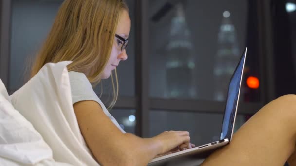 Jovem mulher tendo uma conversa usando um laptop sentado em sua cama com silhuetas de arranha-céus em um backgound — Vídeo de Stock