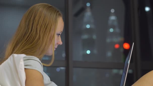 Young woman having a conversation using a laptop sitting on her bed with silhouettes of skyscrappers at a backgound — Stock Video