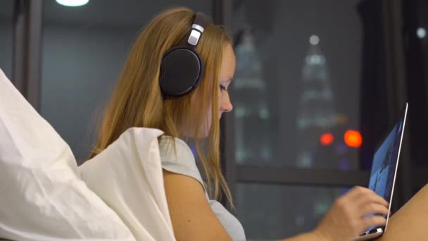 Young woman wearing wireless headphones working using a laptop sitting on her bed with silhouettes of skyscrappers at a backgound — Stock Video